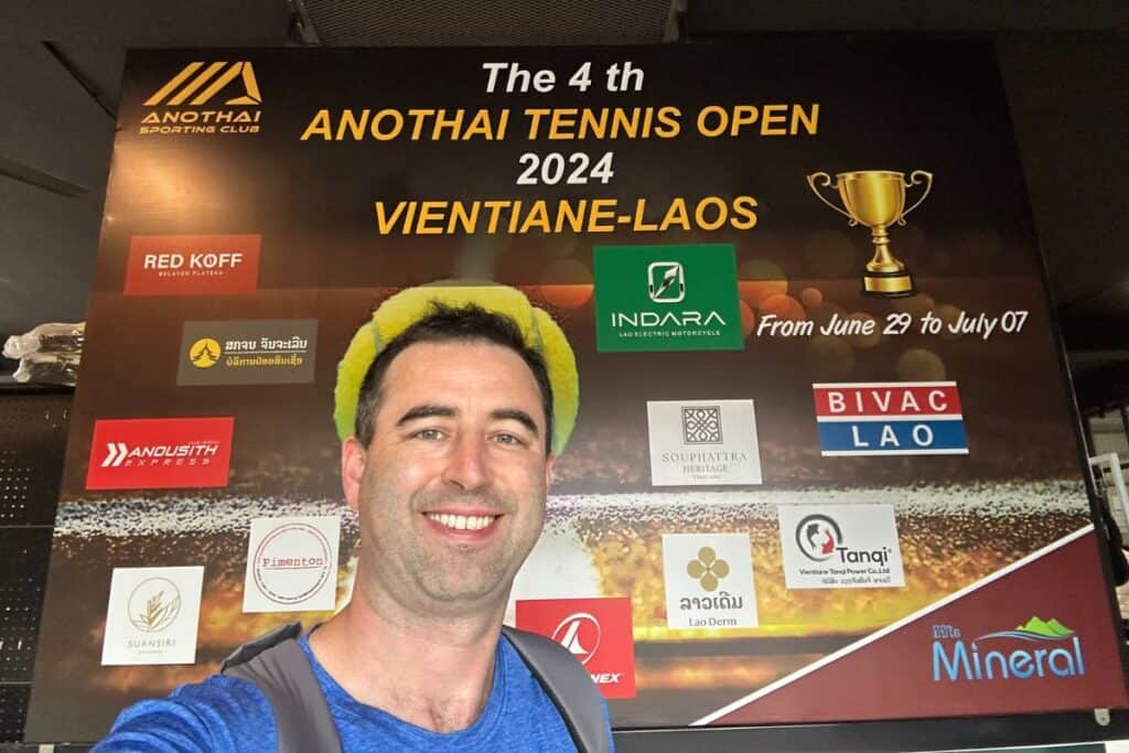 American taking a selfie at Anothai tennis clubhouse, with sign from their tennis tournament, Anothai Tennis Open 2024 Vientiane-Laos.